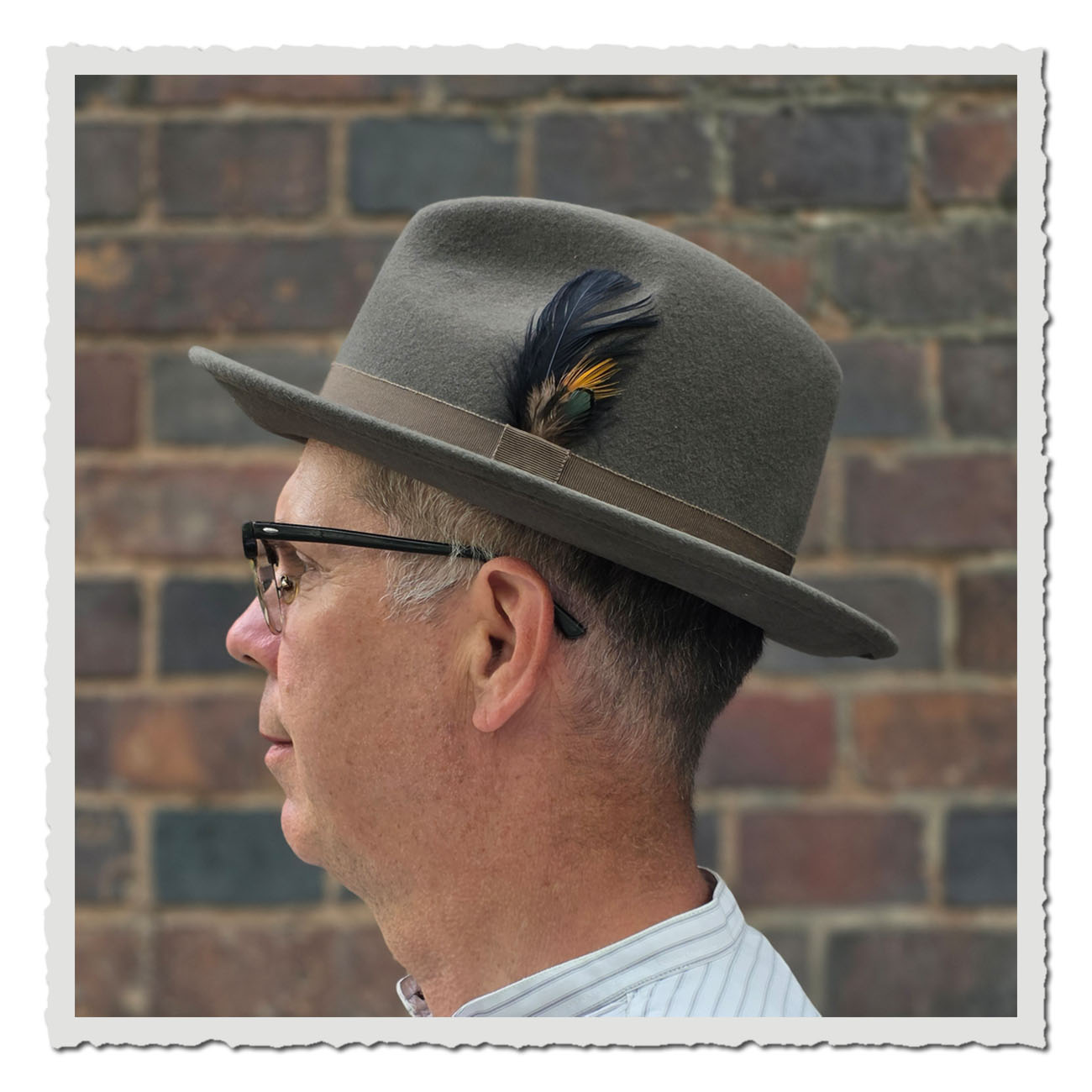 Seitenansicht eines Fedora-Hutes aus grauem Wollfilz, getragen von einem Mann vor einer Backsteinwand. Der Hut hat eine gerade Krempe, ein braunes Ripsband und ist seitlich mit einer dekorativen Feder in Schwarz, Gelb und Grün verziert.