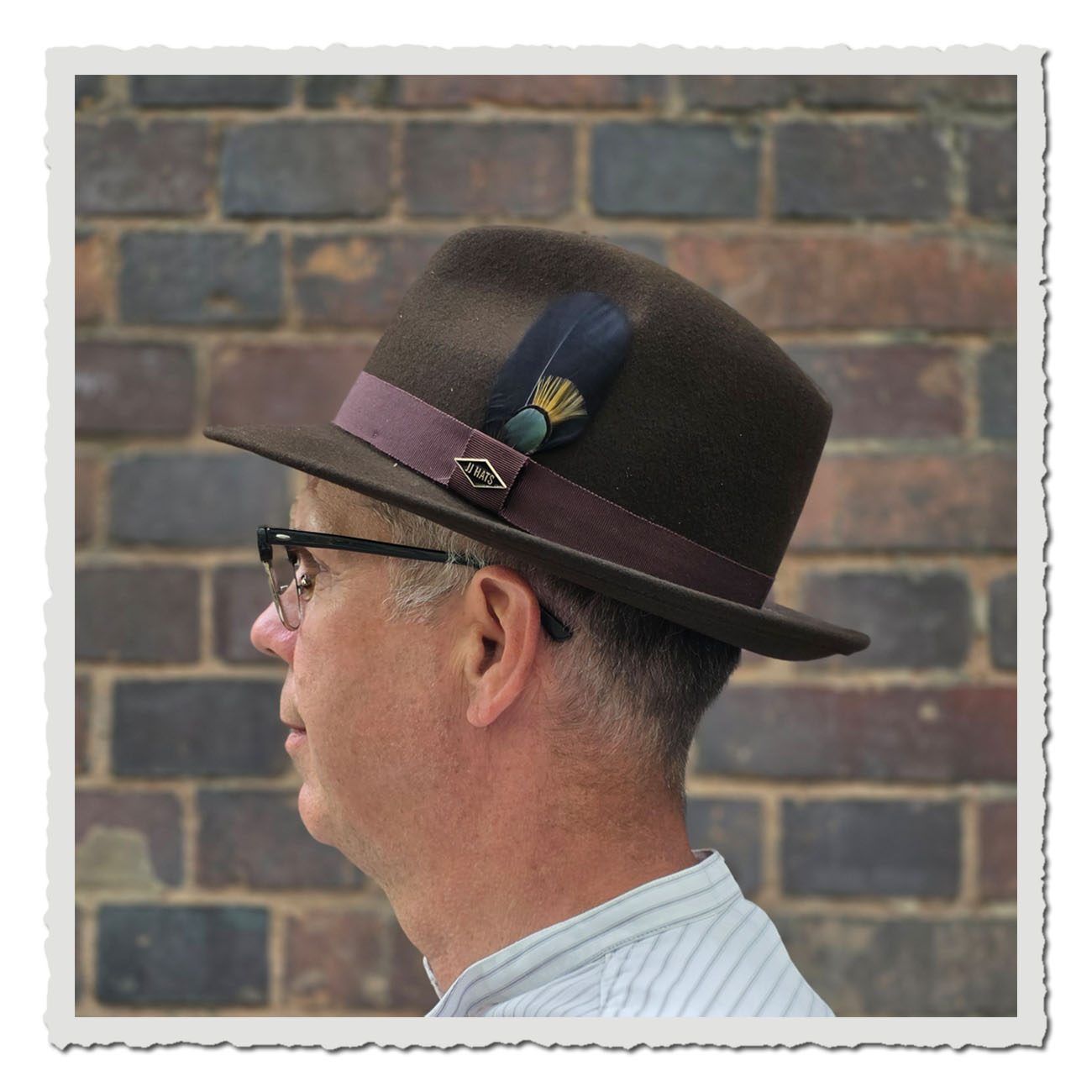 Seitenansicht des Fedora-Hutes Lite Felt vom US Label JJ Hats aus schokobraunem Wollfilz, getragen von einem Mann vor einer Backsteinwand. Der Hut hat eine gerade Krempe, ein weinrotes Ripsband, ein Metallogo-Button und eine dekorative Feder in Schwarz, G