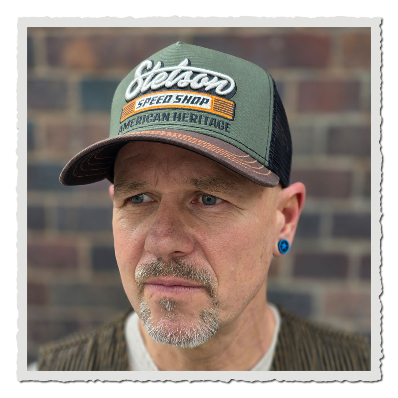 Mann trägt grün-braunes Stetson Trucker Cap mit Stickerei „Speed Shop American Heritage“.