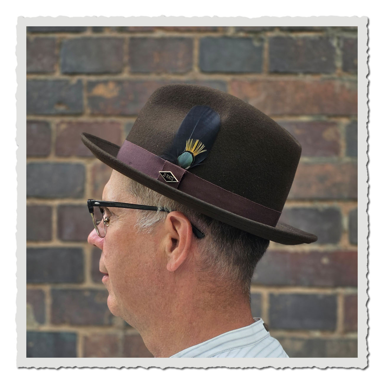 Seitenansicht vom Fedora-Hutes Lite Felt von der US Marke JJ Hats aus braunem Wollfilz, getragen von einem Mann vor einer Backsteinwand. Der Hut hat eine gerade Krempe, ein weinrotes Ripsband und eine dekorative Feder in Schwarz, Grün und Gelb.